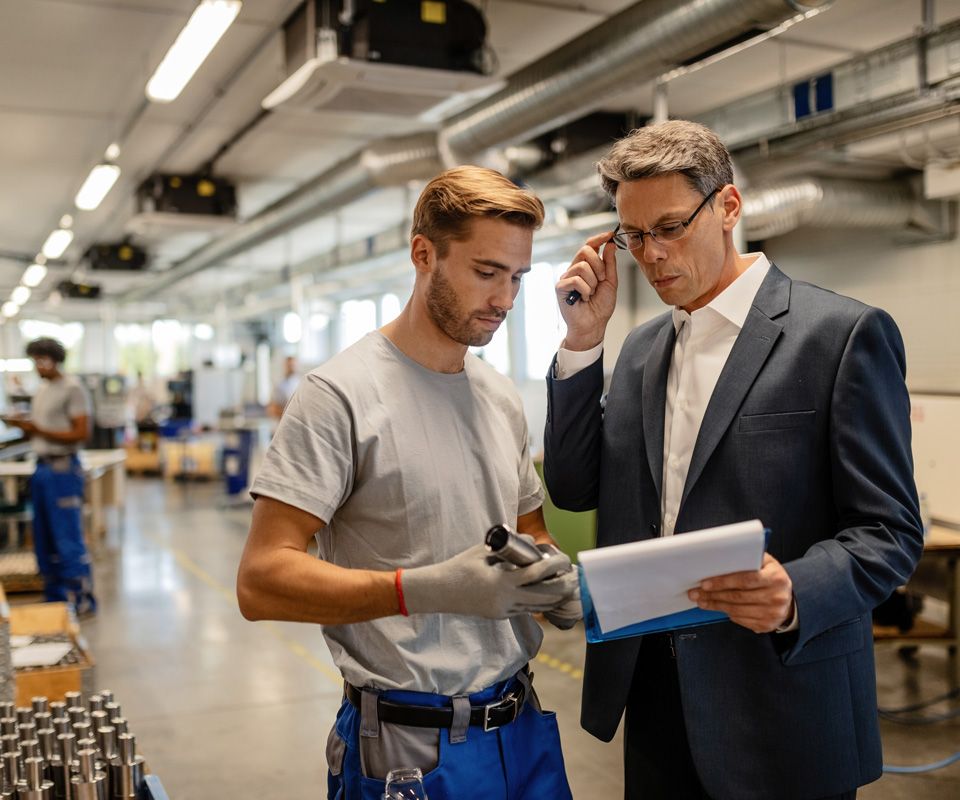 Two men talking on a and reviewing a document inside a factory setting.